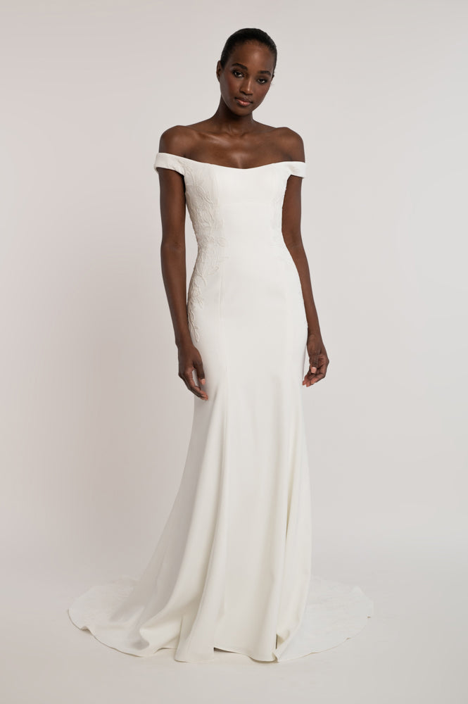 An off-the-shoulder, floor-length white wedding gown with a fitted bodice and soft train, shown on a model against a plain light gray studio background.