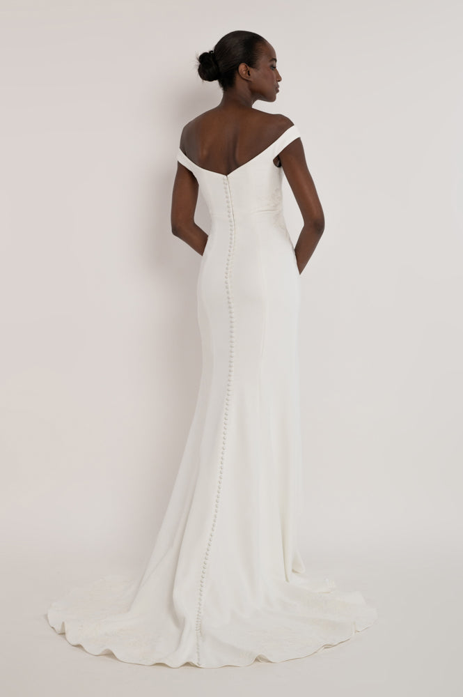 A woman modeling an off-the-shoulder white wedding gown from the back, showing a fitted silhouette, buttoned back, and a long train against a plain light background.
