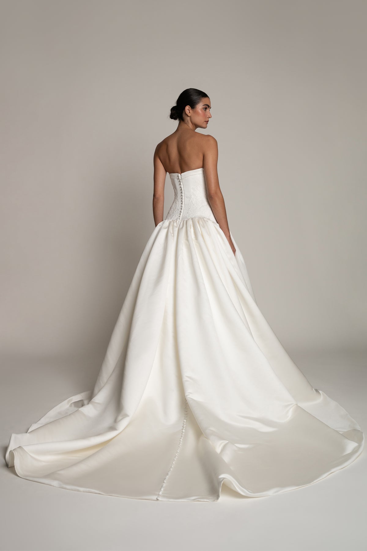 A woman modeling a strapless white wedding gown from the back, showing a structured corset-style bodice and a full satin train, against a plain warm beige studio background.