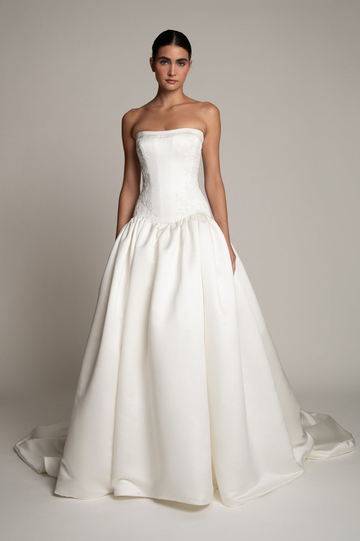 A woman modeling a strapless white wedding gown with a structured bodice, full satin skirt, and train against a plain light gray studio background.