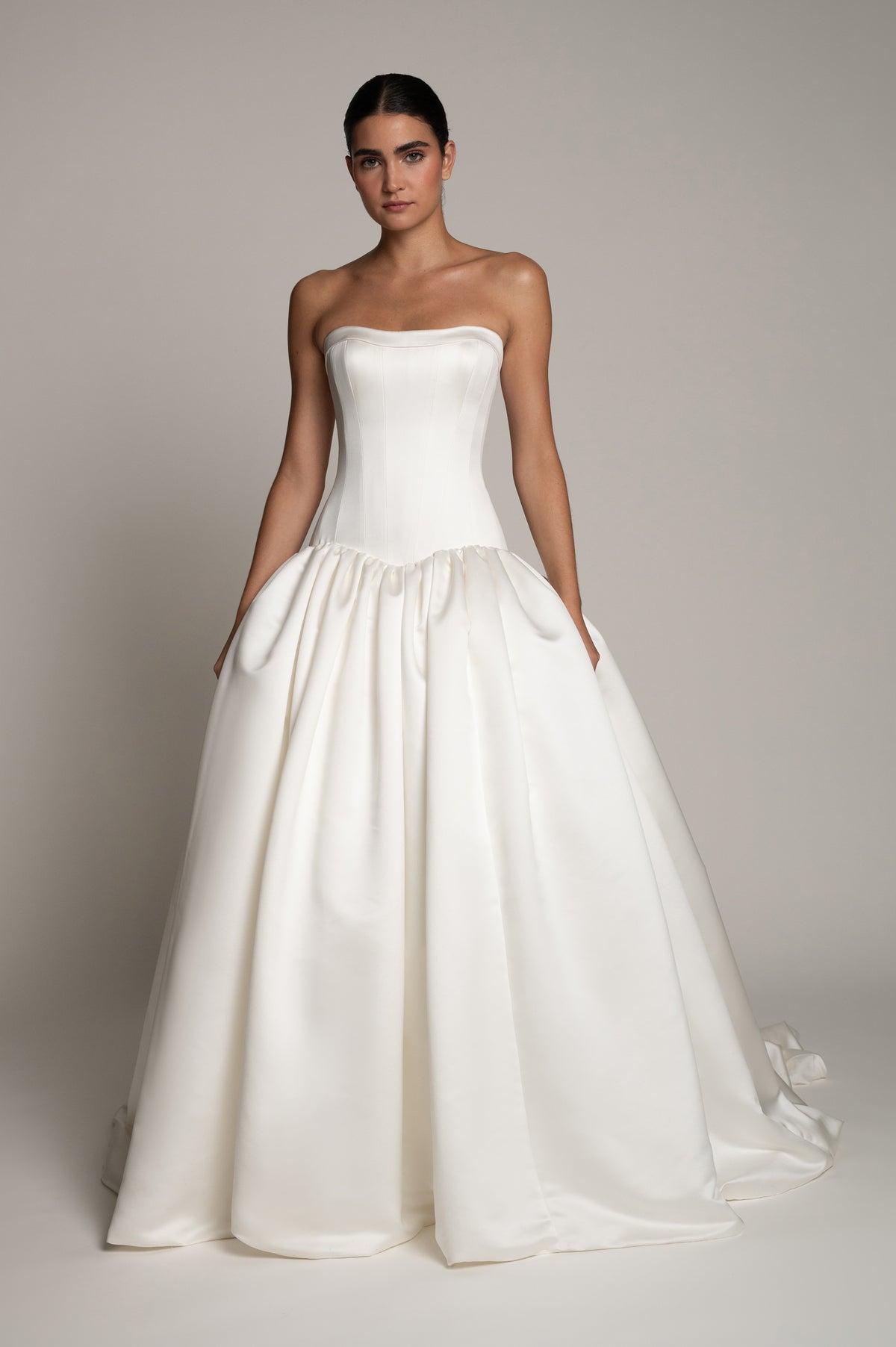 A woman modeling a strapless white ball gown wedding dress with a structured corset bodice and a voluminous satin skirt, photographed against a plain light gray studio background.