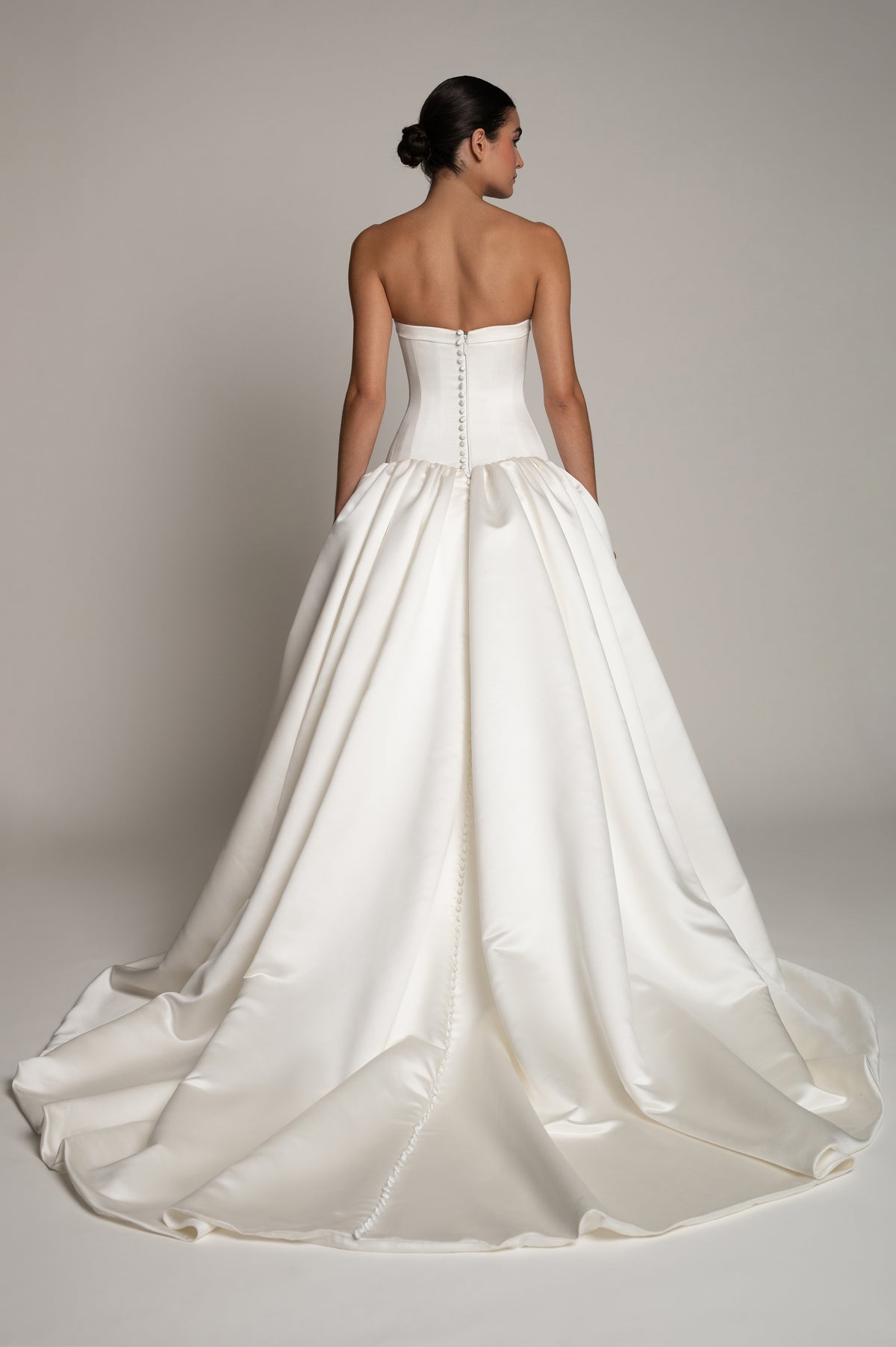 A strapless white satin wedding gown shown from the back on a model, featuring a structured corset-style bodice with button detailing and a full, flowing skirt with a train, set against a neutral gray studio background.