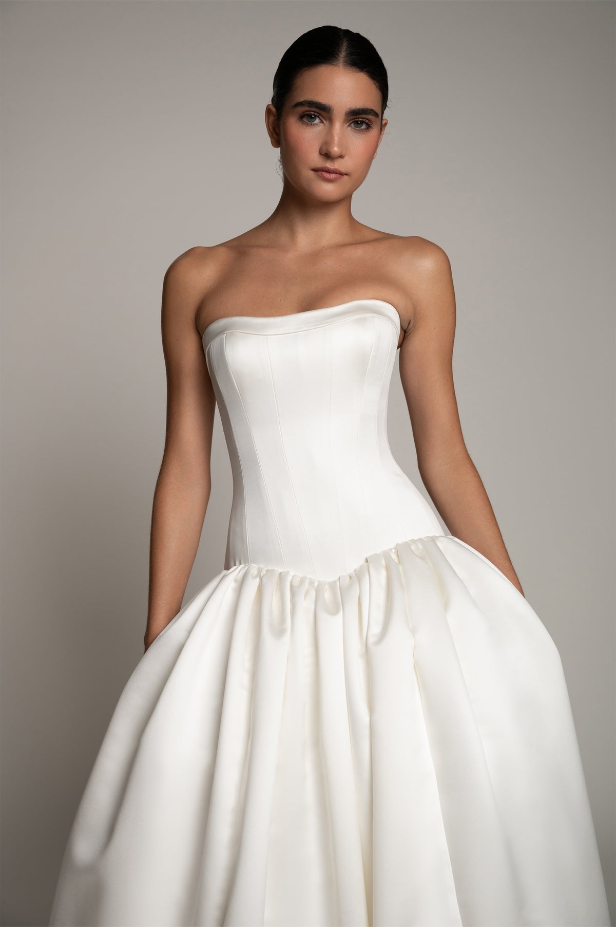 A woman modeling a strapless white satin wedding gown with a structured corset-style bodice and a voluminous gathered skirt, photographed against a plain light gray studio background.