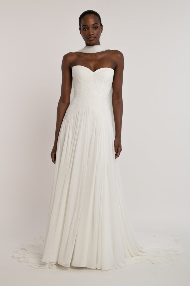 A woman modeling a strapless white wedding gown with a sweetheart neckline, ruched bodice, and flowing pleated skirt against a plain light background.