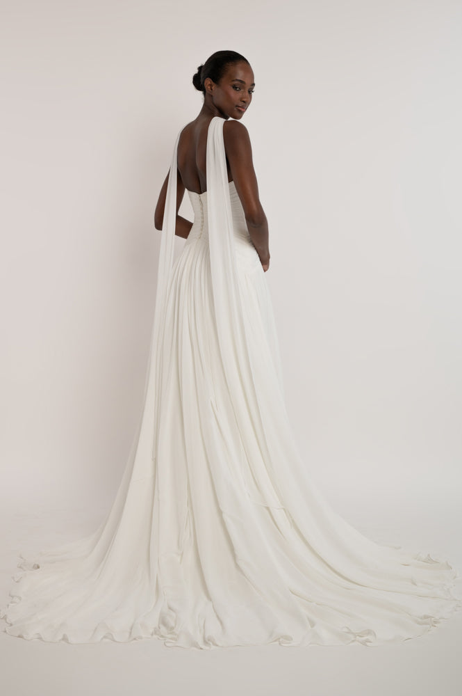 A model from behind wearing a flowing white bridal gown with a long train, photographed against a plain light background.