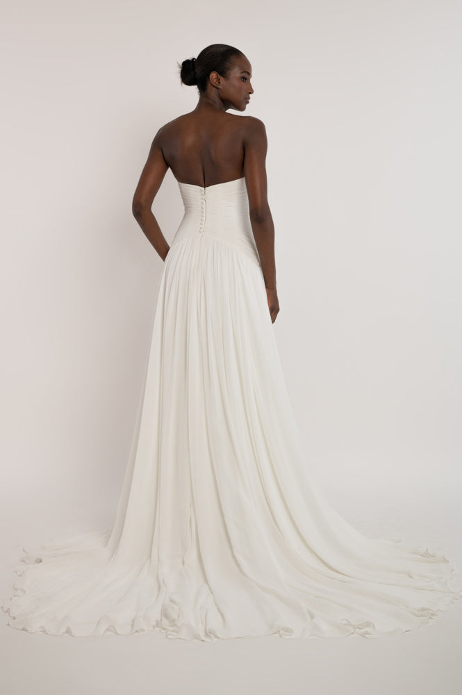 A woman modeling a strapless white wedding gown from the back, showing a structured corset-style bodice with a long flowing skirt and train against a plain light background.