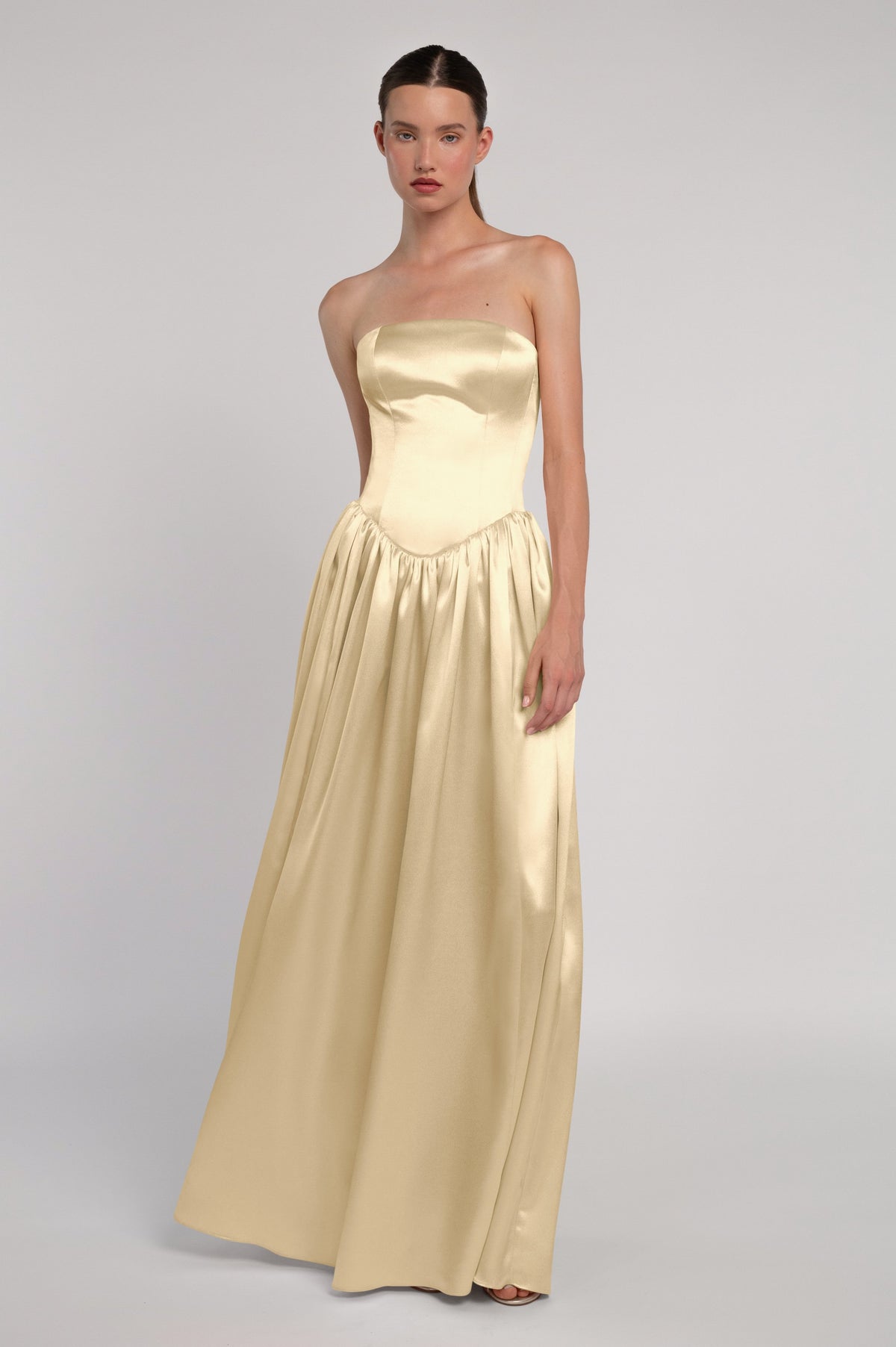 A model wearing a strapless champagne-gold satin maxi dress with a structured bodice and flowing skirt, photographed against a neutral light gray studio background.