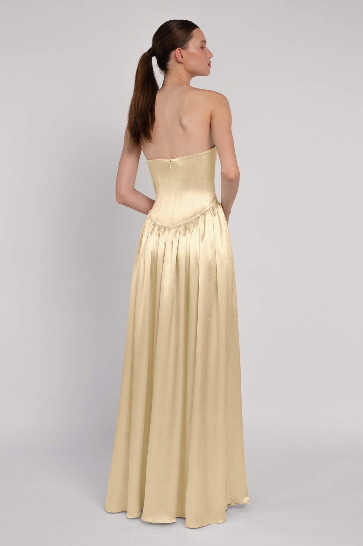 A woman modeling a strapless floor-length gown from the back, shown against a plain light gray studio background. The dress has a structured corset-style bodice and a flowing satin-like skirt in a warm pale gold color.