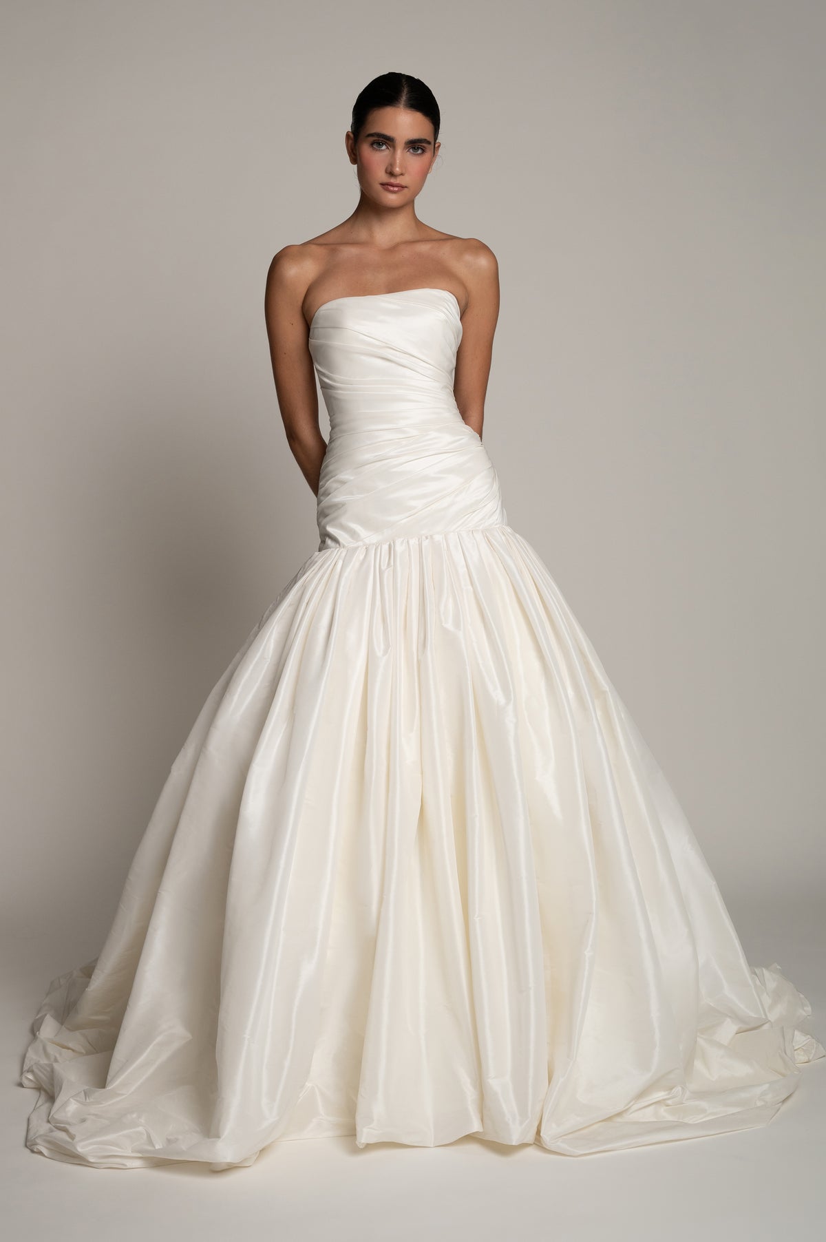 A woman modeling a strapless white ball gown wedding dress with a ruched bodice and a full, flowing skirt against a plain light gray studio background.