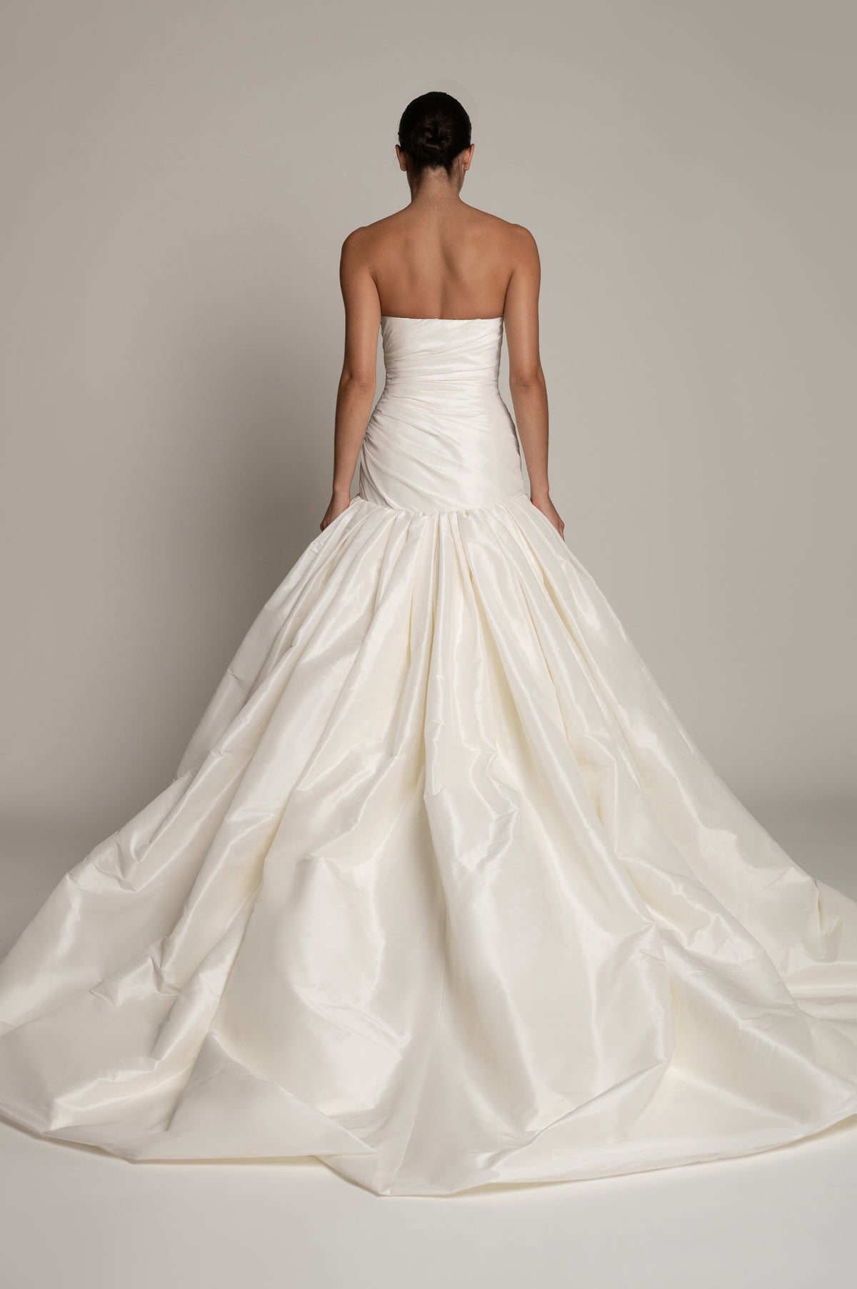 A strapless white taffeta wedding gown shown from the back on a model, featuring a structured ruched bodice and a voluminous, draped ball gown skirt against a neutral studio backdrop.