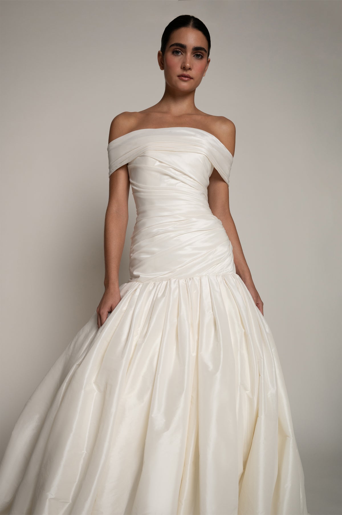 An off-the-shoulder white taffeta wedding gown with a ruched bodice and a voluminous, pleated skirt, shown on a model against a neutral studio background.