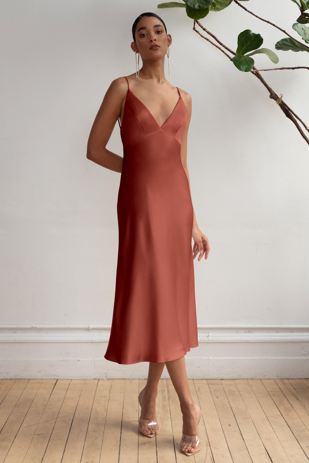 Woman wearing a brown slip dress standing against a white wall with a plant.