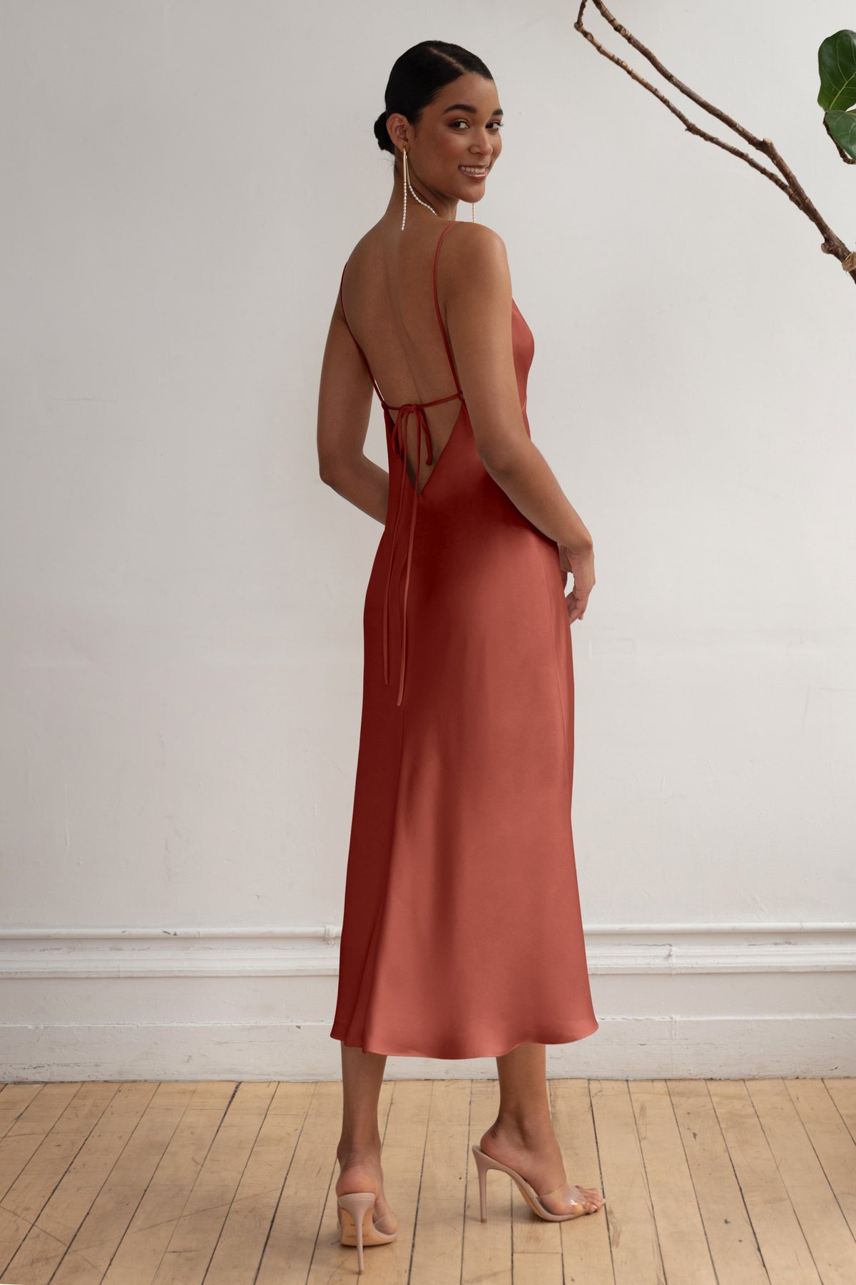 Woman wearing a rust-colored dress with a backless design, standing against a white wall.