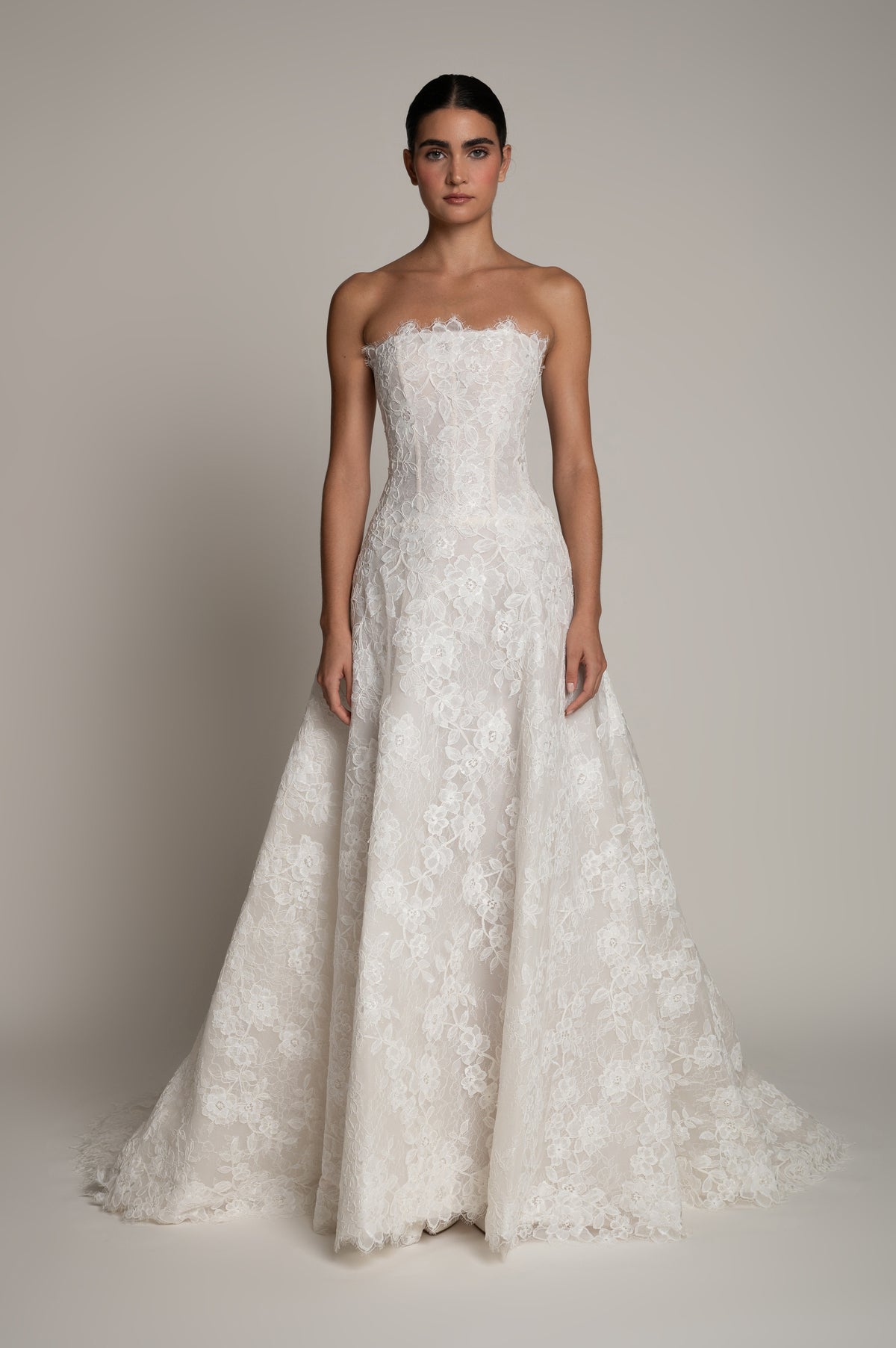 A strapless white lace wedding gown with a fitted bodice and full flared skirt, shown on a model against a plain warm gray studio background.