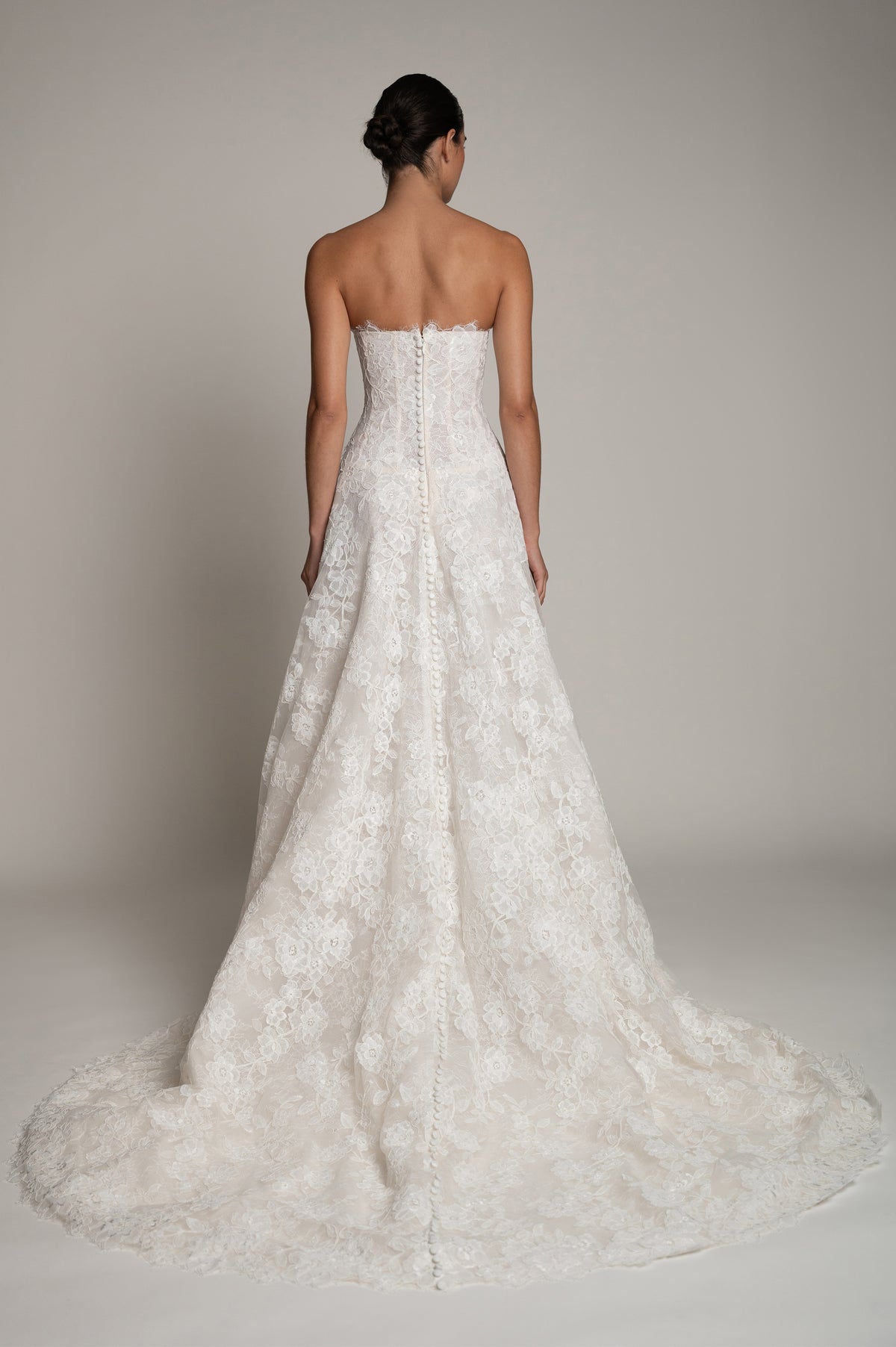 A strapless white lace wedding gown shown from the back on a model, featuring a fitted bodice, lace overlay, and a full flared skirt with a long train against a neutral studio background.