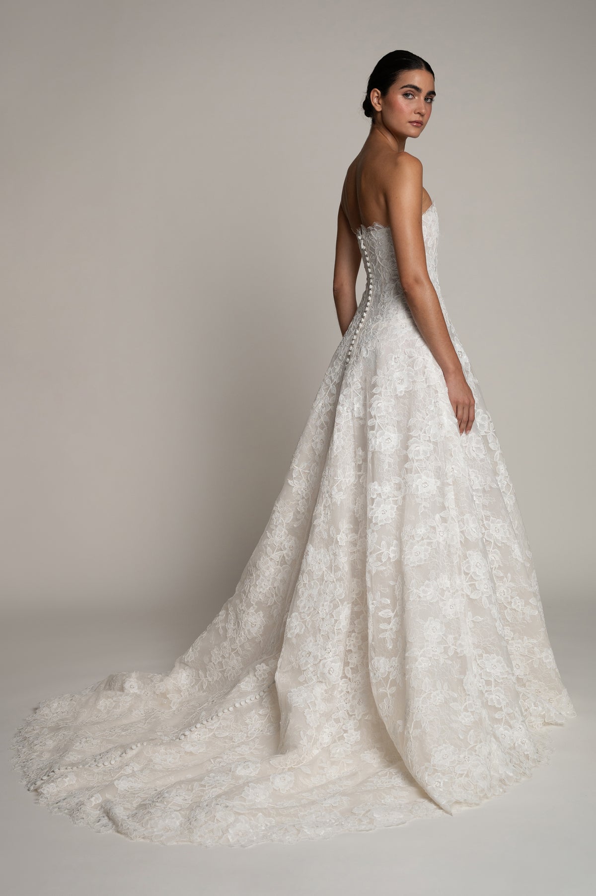 A woman modeling a strapless white lace wedding gown with a fitted bodice, full flared skirt, and long train against a plain warm beige studio background.