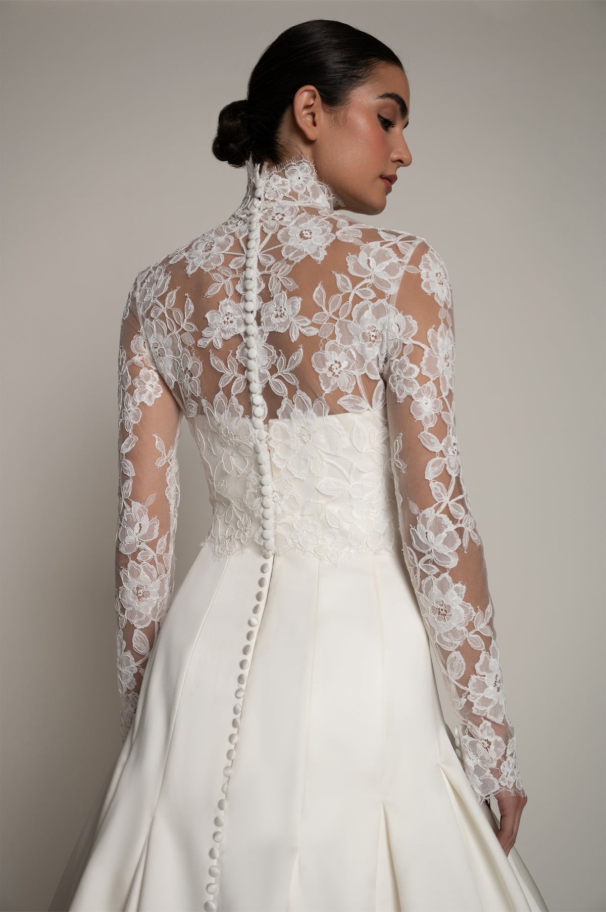 A woman shown from the back wearing a white bridal gown with a sheer floral lace high-neck long-sleeve back, a fitted lace-embroidered bodice, and a full skirt, photographed against a plain light gray studio background.