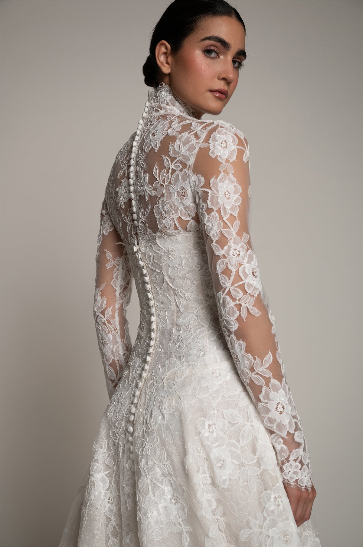 A woman modeling a white lace bridal gown from the back, showing long sheer sleeves, floral lace detailing, and a beaded down the spine, against a neutral gray studio background.