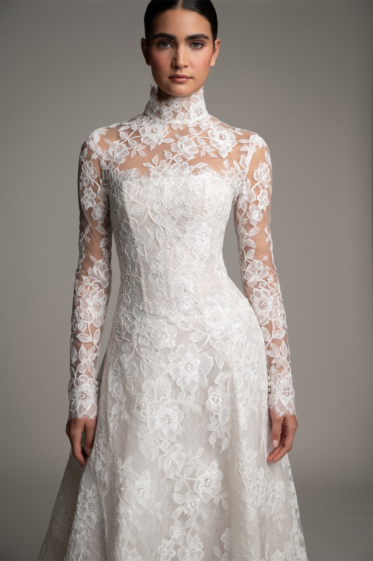 A woman modeling a long-sleeve high-neck white lace wedding dress against a plain gray studio background.