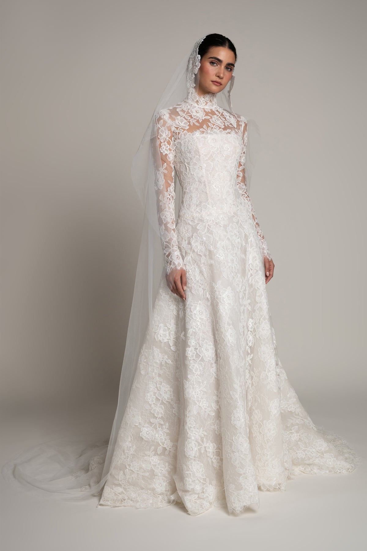 A woman modeling a long-sleeve lace wedding gown with a high mock neck, fitted bodice, and full skirt, wearing a sheer veil against a plain light gray studio background.