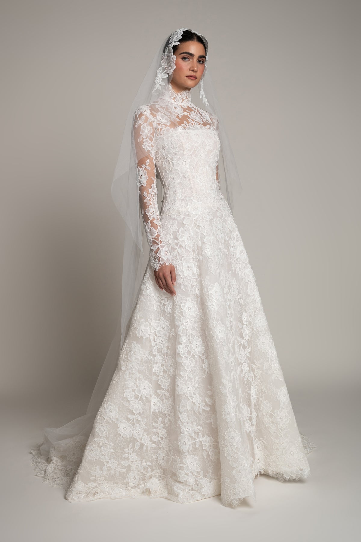 A woman modeling a white lace wedding gown with long sheer lace sleeves, a high neckline, and a long sheer veil against a plain light gray studio background.