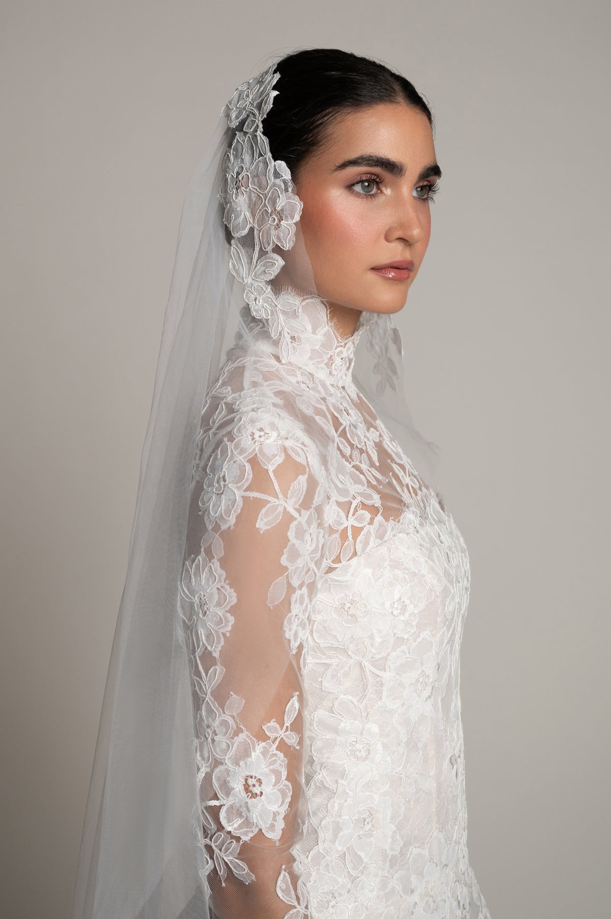 A woman in profile wearing a white lace bridal gown with floral lace sleeves, a matching sheer veil with lace flower accents, and dark hair styled back, photographed against a plain light gray studio background.