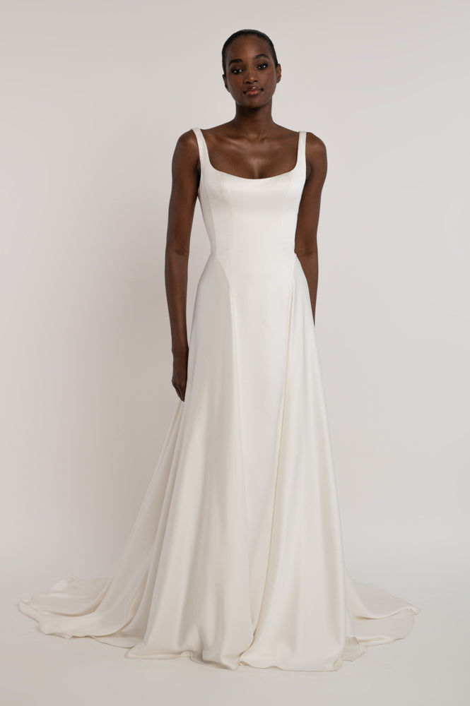 A woman modeling a sleeveless white A-line wedding gown with a square neckline and a soft train against a plain light gray studio background.
