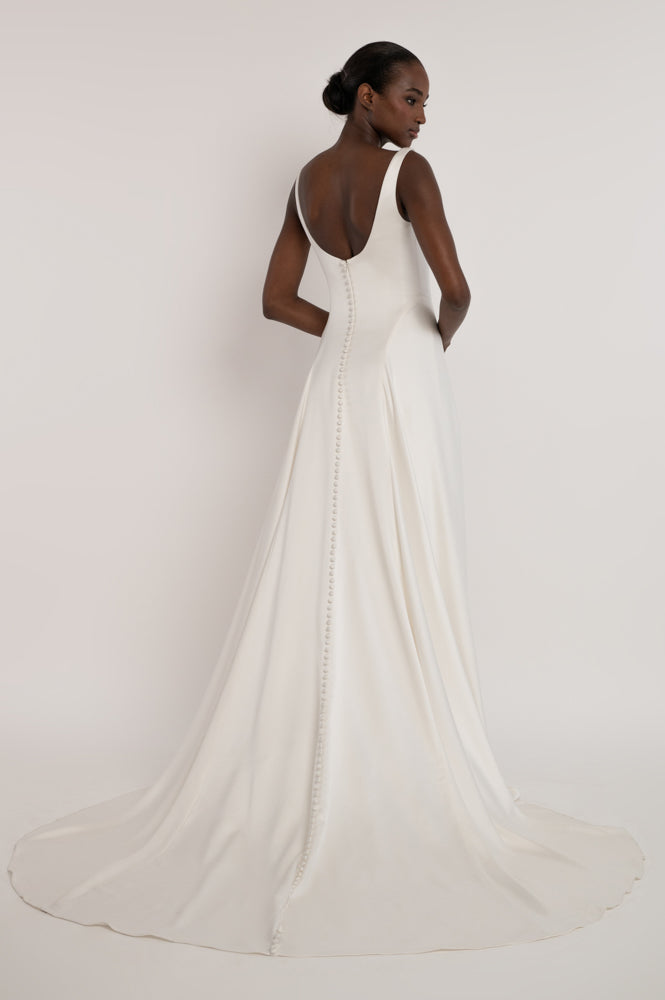 A model shown from the back wearing a white sleeveless wedding dress with a deep V back, buttoned center seam, and a long flowing train against a plain light background.