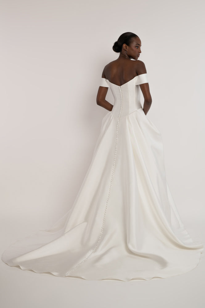A woman modeling an off-the-shoulder white wedding gown from the back, showing a structured corset-style bodice and a full satin train against a plain light background.