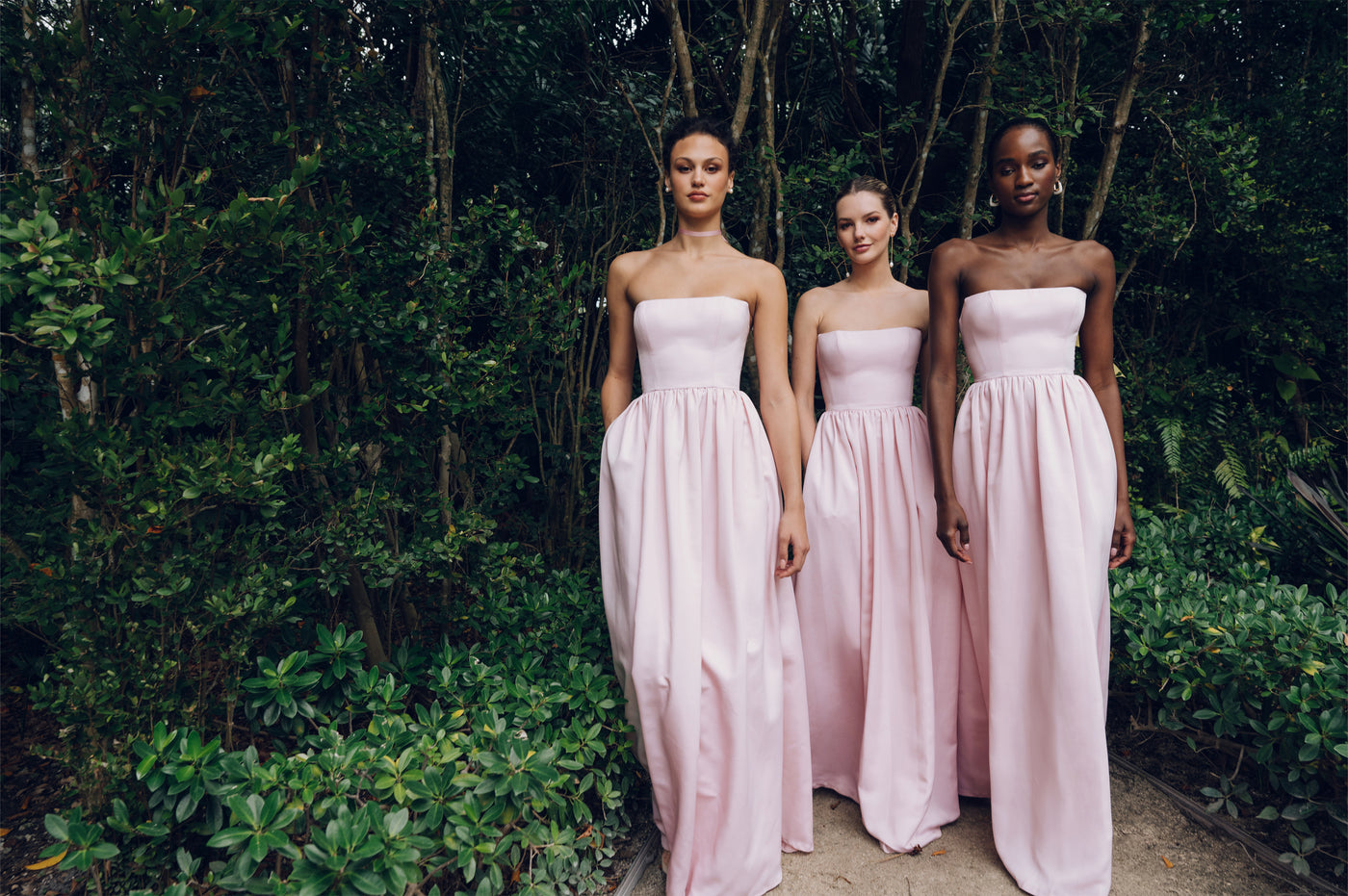 Three bridesmaids wearing light pale powder pink Faille bridal party dresses in outdoor setting with greenery.