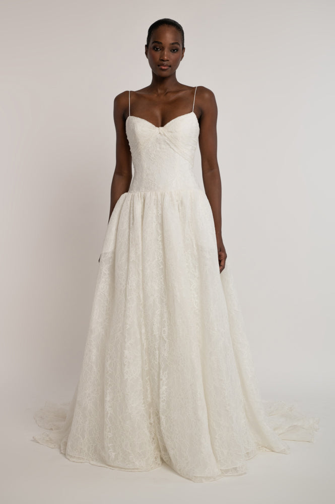 A model wearing a white lace wedding gown with thin spaghetti straps, a sweetheart neckline, and a full A-line skirt, photographed against a neutral light gray studio background.