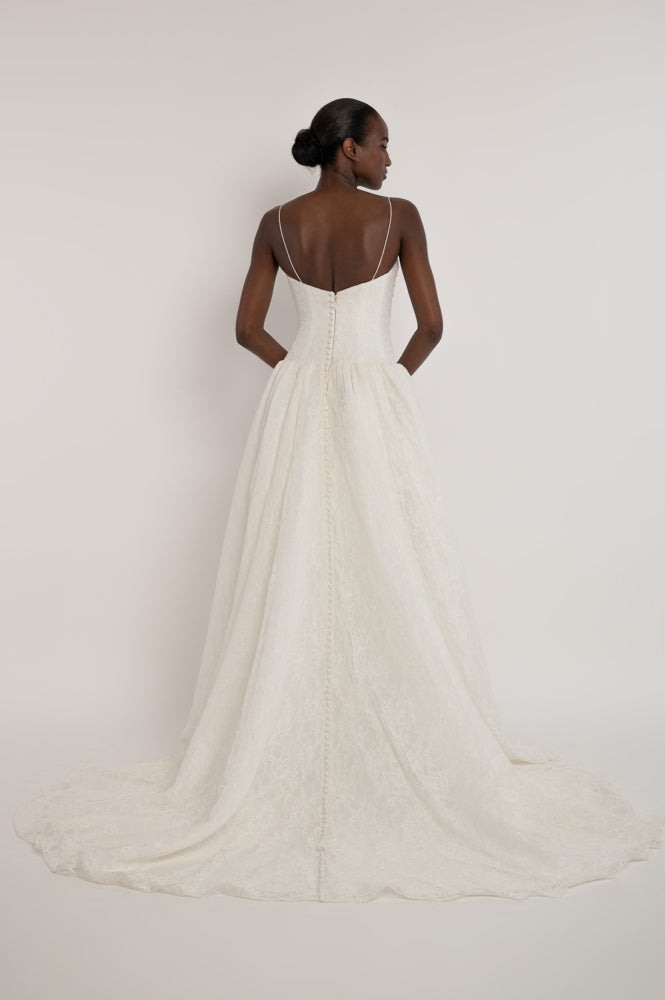 A model shown from the back wearing a white wedding dress with thin spaghetti straps, a fitted bodice, and a voluminous lace-trimmed skirt with a long train, set against a plain off-white studio background.