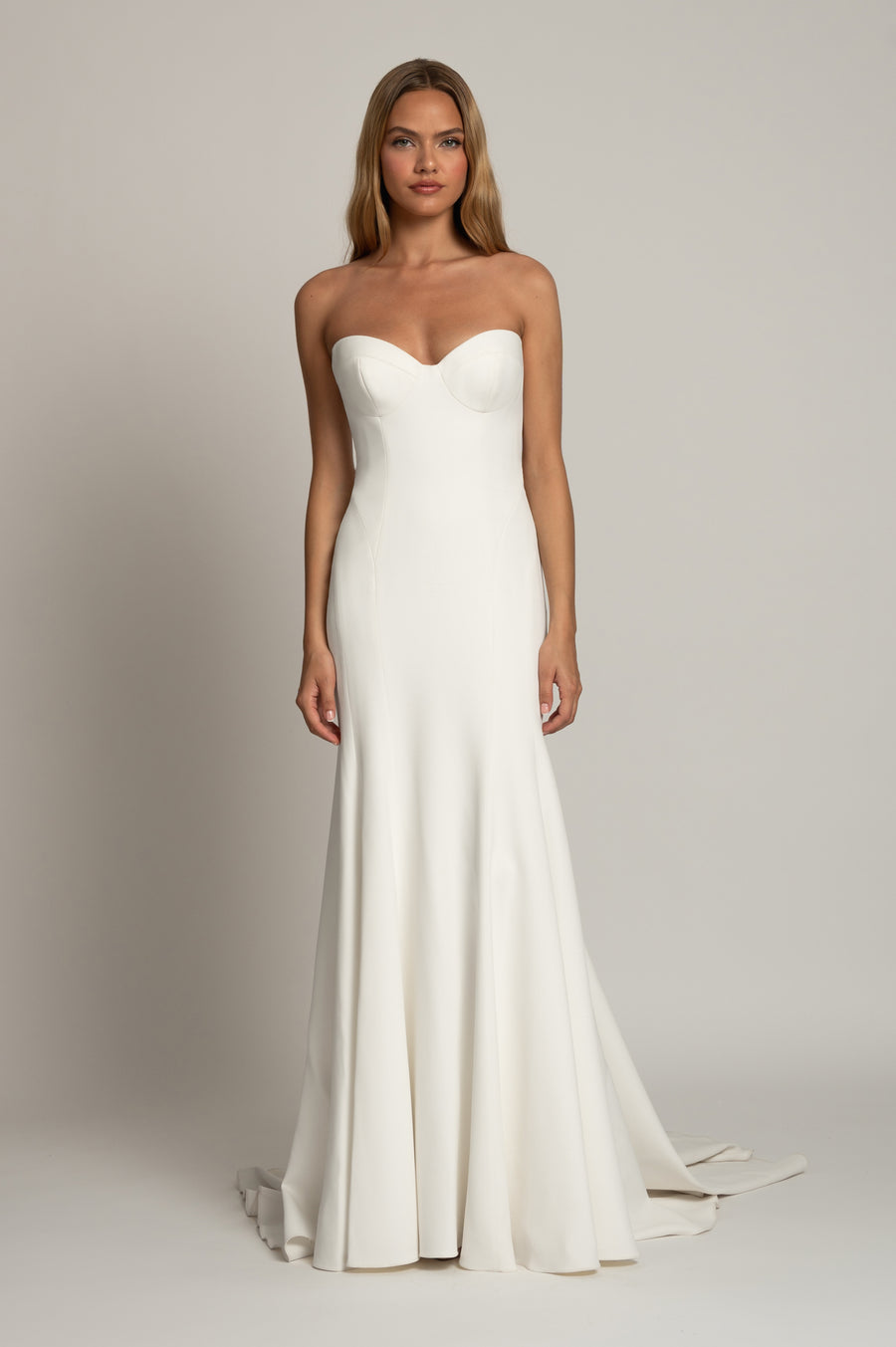 A woman modeling a strapless white wedding gown with a sweetheart neckline, fitted bodice, and floor-length skirt with a slight train, photographed against a plain light gray studio background.