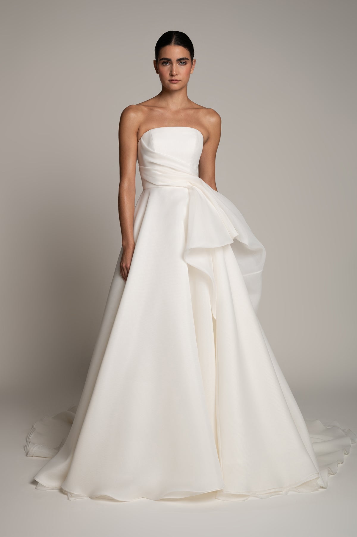 A woman modeling a strapless white wedding gown with a structured bodice, draped waist detail, and a full layered skirt against a plain warm gray studio background.