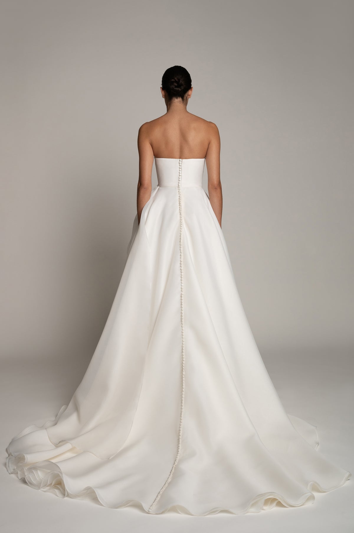 A woman shown from the back wearing a strapless white wedding gown with a structured bodice and a long flowing train against a plain light gray studio background.