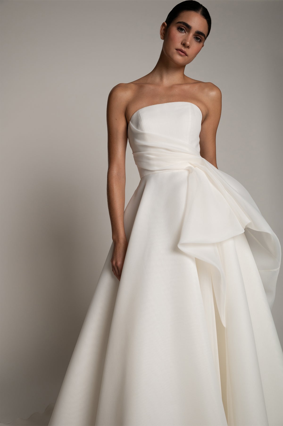 A woman modeling a strapless white wedding gown with a gathered, layered skirt against a plain light gray studio background.