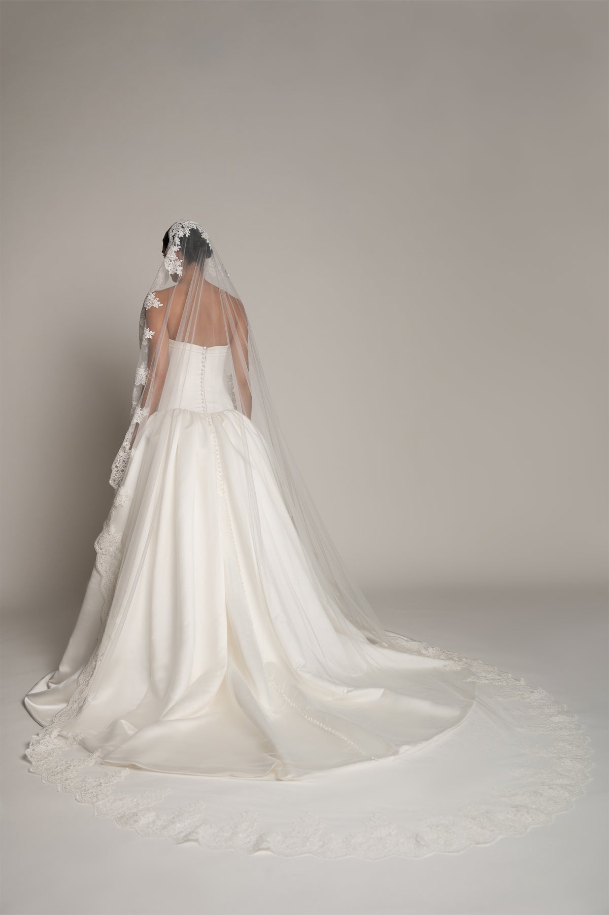 A woman wearing a strapless white wedding gown with a long sheer veil, photographed from the back against a plain light gray studio background.