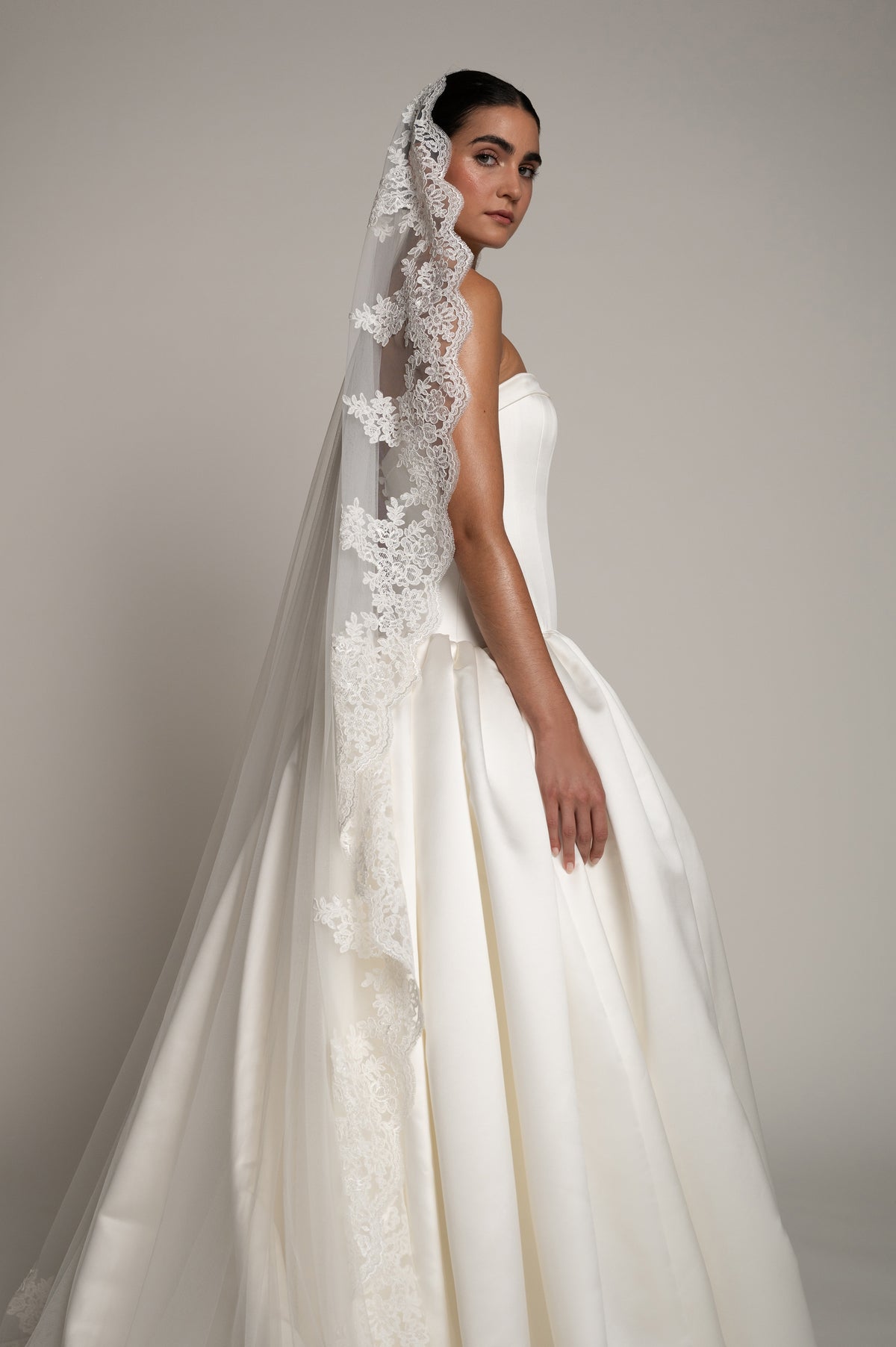 A woman modeling a strapless white wedding gown with a long sheer veil featuring floral lace applique, photographed against a plain warm gray studio background.