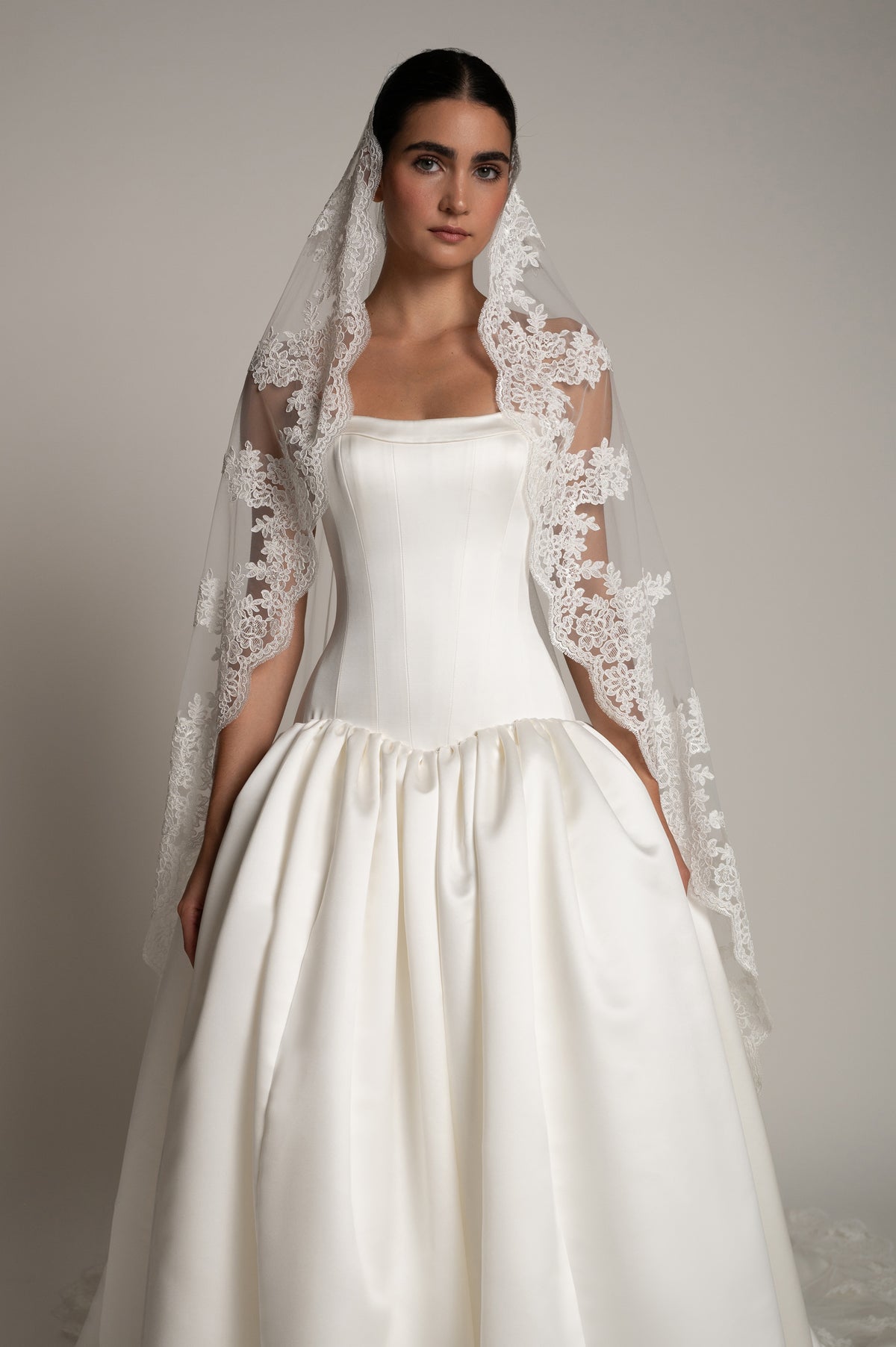 A woman modeling a white wedding dress with a sheer lace-trimmed veil, photographed against a plain light gray studio background.