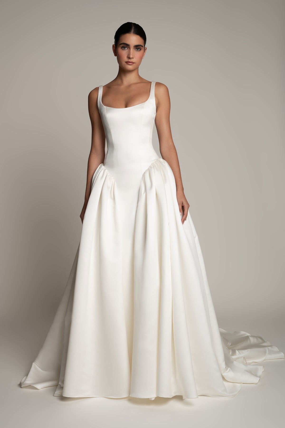 A woman modeling a sleeveless white wedding gown with a structured bodice, full gathered skirt, and a long train against a plain warm gray studio background.