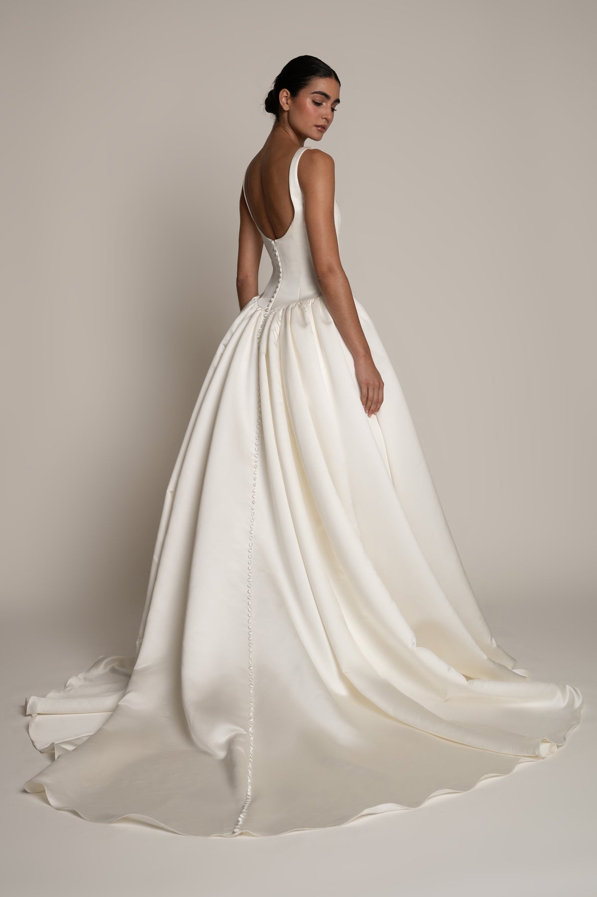 A woman modeling a bridal gown from the back, showing a structured ivory satin skirt with a long train and a low open back, against a plain warm beige studio background.