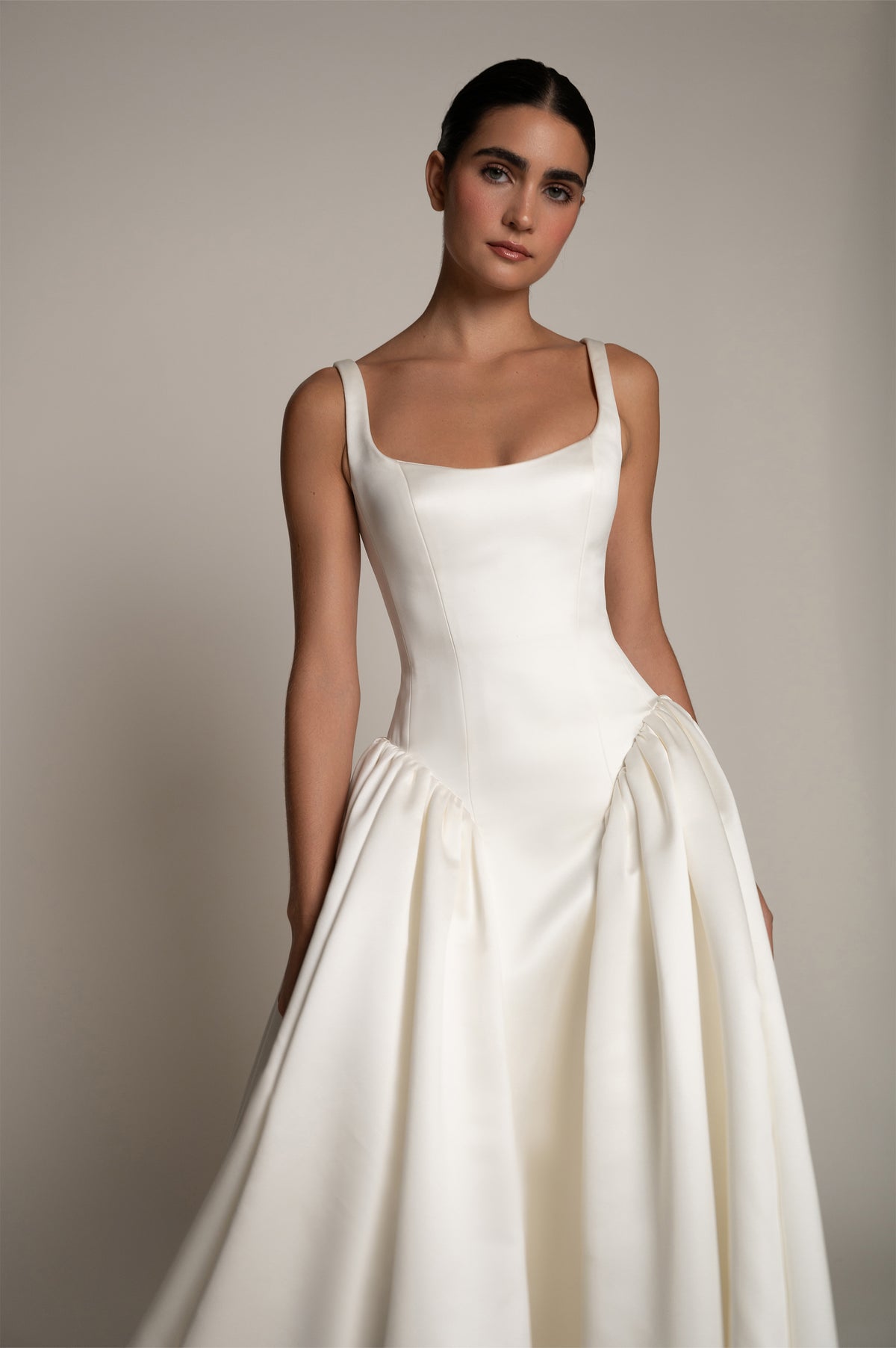 A woman modeling a white sleeveless wedding dress with a structured bodice, square neckline, and full gathered skirt against a plain warm gray studio background.