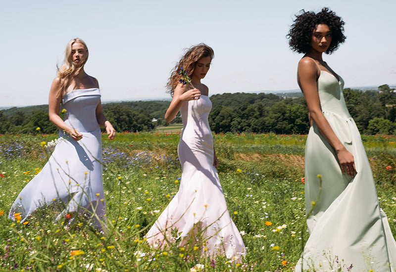 Three women wearing floor-length strapless bridesmaid dresses in soft pastel shades of lavender, blush, and sage green, walking through a sunlit wildflower meadow.