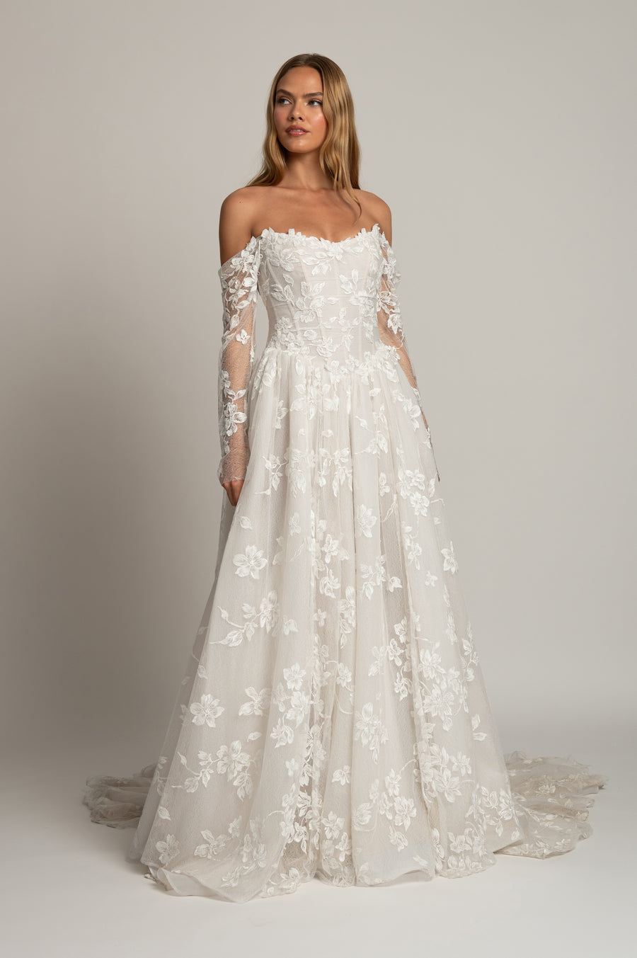An off-the-shoulder A-line wedding gown with long sheer lace sleeves and a full skirt covered in floral lace applique, shown on a model against a neutral studio background.