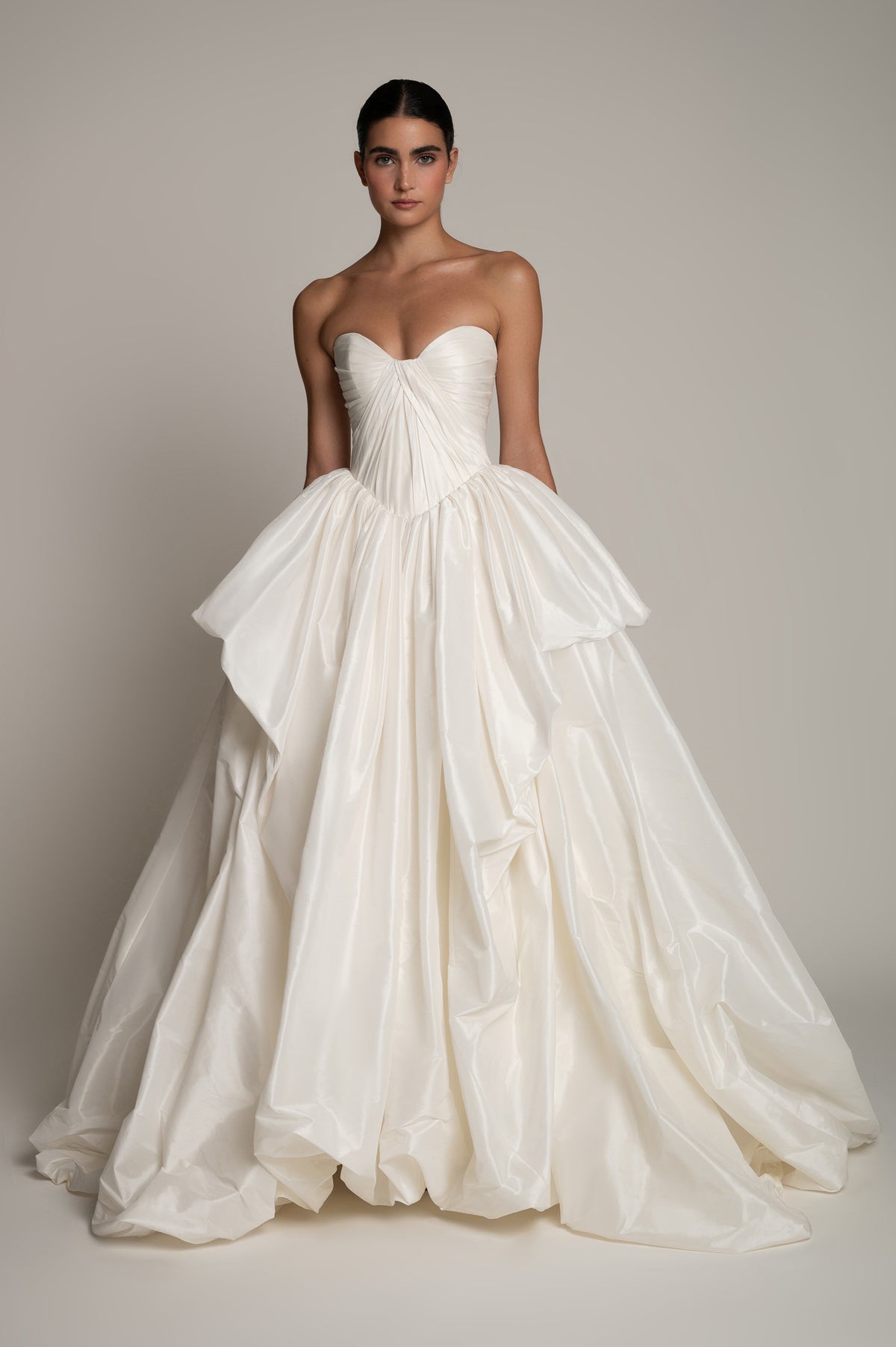 A model wearing a strapless white taffeta wedding gown with a structured sweetheart bodice, gathered corset waist, and a voluminous layered ruffle skirt, photographed against a neutral warm gray studio backdrop.