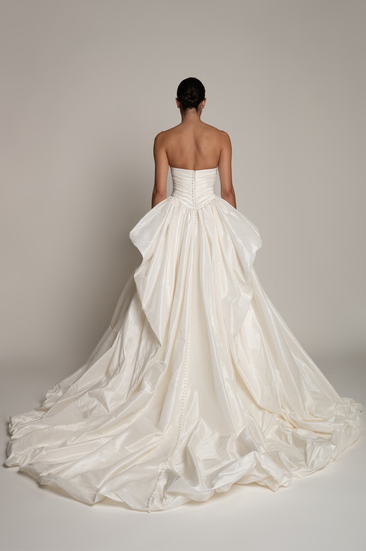 A woman viewed from behind wearing a strapless white bridal gown with a structured corset-style bodice and a voluminous, layered tulle skirt that spreads across the floor against a plain light gray studio background.