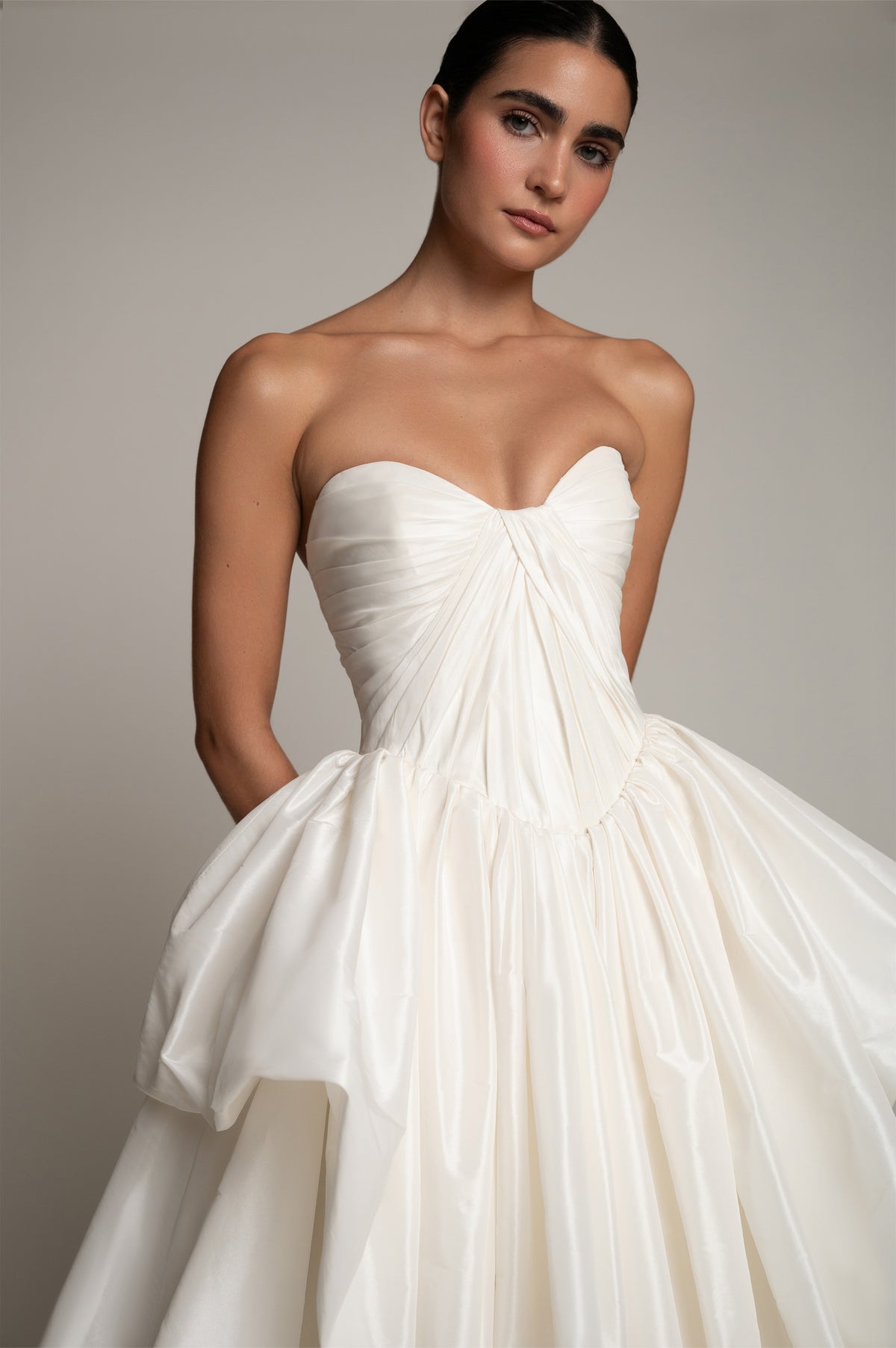 A woman modeling a strapless white wedding gown with a sweetheart neckline, ruched bodice, and voluminous gathered skirt against a plain light gray studio background.